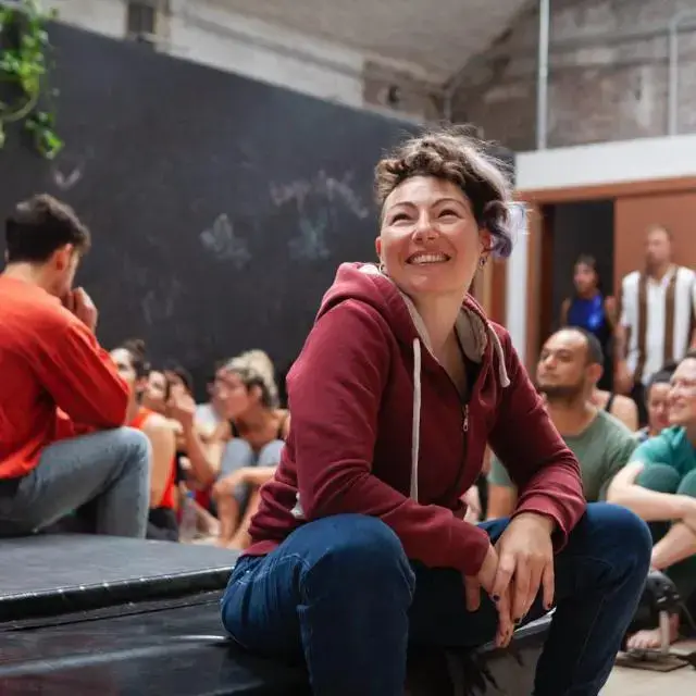 irene-cecotti-circus-passion-profile Profile picture of Irene Cecotti smiling joyfully, with short hair and a maroon hoodie, seated on a dark performance platform during a circus practice session, surrounded by other blurred participants in a well-lit studio setting.